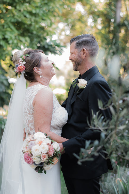 bride and groom laughing with a beautiful floral head piece , bouquet and button hole created by mudgee monkey florist in shades of pink and peach