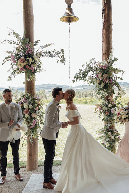 wedding couple man and lady in wedding outfits exchange a first kiss at The pavilon at lowes bush chapel with flowers on the arbour by Mudgee monkey florist. At lowes wines