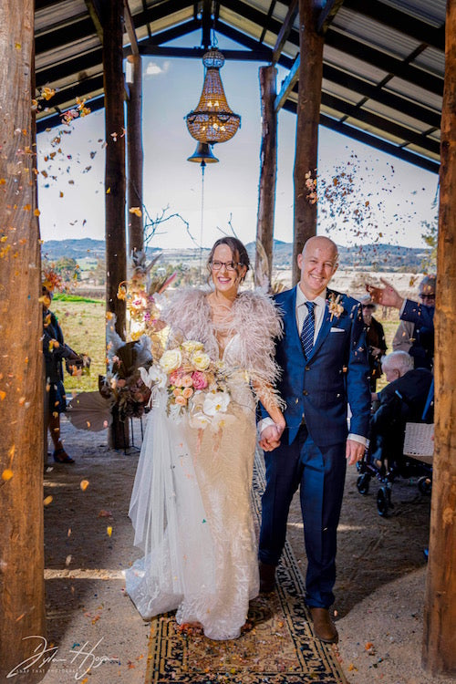 bride and groom walking out of the lowes bush chapel amonst a petal toss of pink and peach rose petals. Bouquet of roses and orchids in shades of lemon, pink and peach made by mudgee monkey florist