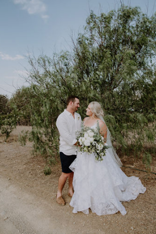 bride and groom in the bush. Groom has shorts and bride in long white dress holding flowers by mudgee monkey florist