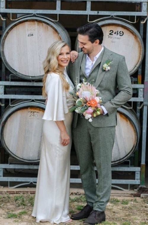 couple in wedding clothes pose next to wine barrels with a king protea and blsuh weedding bouquet and button hole from Mudgee monkey florist 
